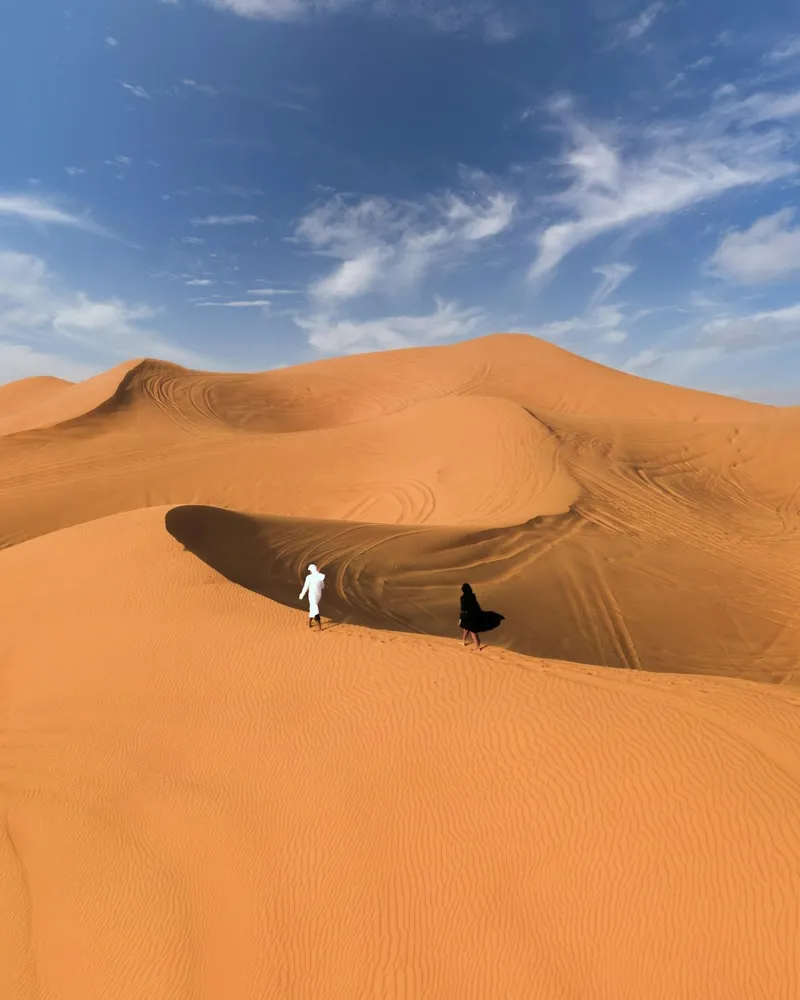 Placeholder: two figures crossing a tall orange dune in the desert outside Dubai
